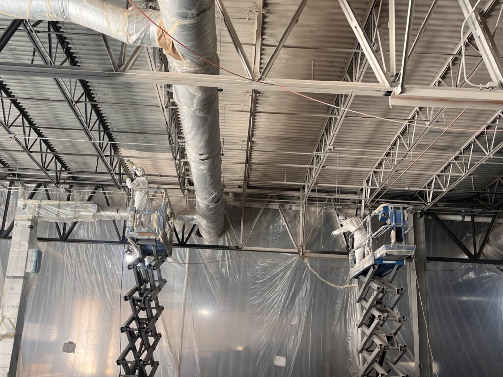 Workers on scissor lifts spraying paint or fireproofing on ceiling structures and ducts for Bell SPI - Shamrock Painting in Denver, CO.