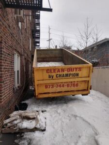 A yellow roll-off dumpster from Champion Waste Removal, Inc. placed in a snowy area next to a building in Orange, NJ.