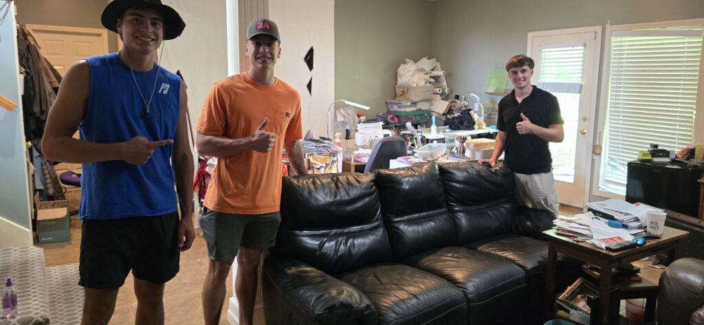 The Young Bucks Junk team standing next to a black leather couch in a cluttered living room in Shawnee, KS, ready for removal.
