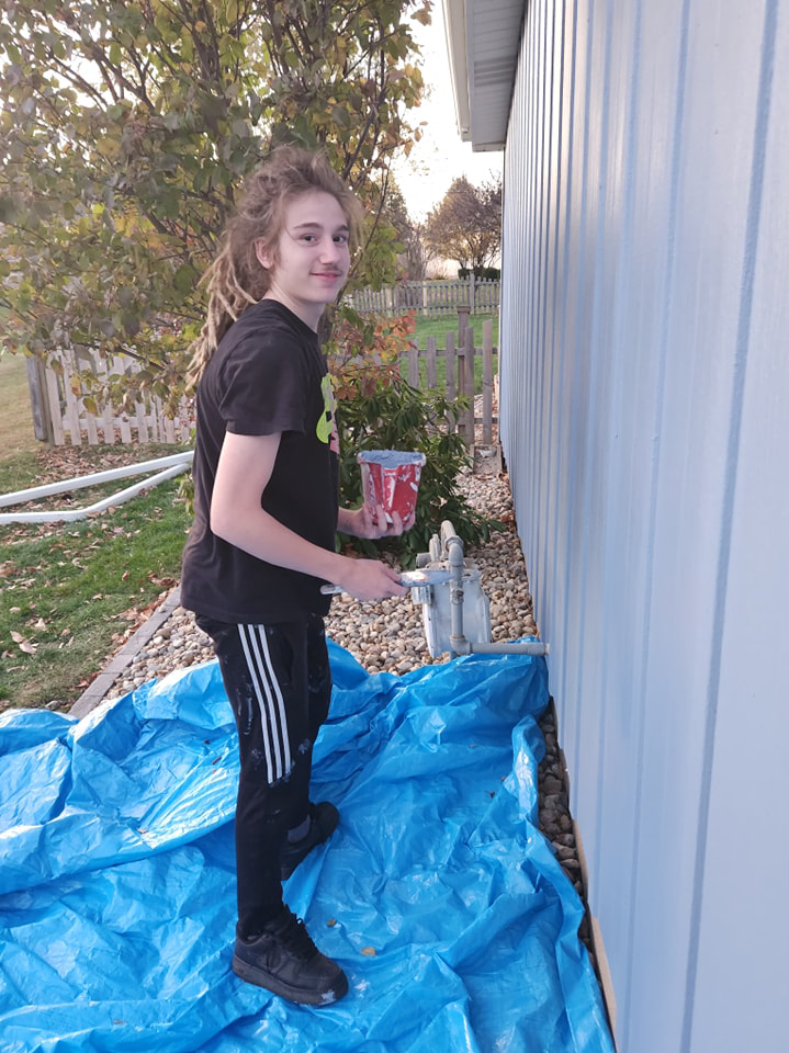 A young man holding a paint can and roller, standing on a blue tarp while painting house siding for Drew's Handy Construction in Charleston, IL.