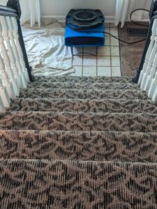 An air mover drying carpeted stairs and adjacent tiled floor after water extraction by R&W Carpet Cleaning & Water Extraction in Independence, MO.