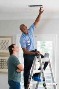 An Aire Serv technician on a ladder servicing an air vent on the ceiling while a customer watches in Billings, MT.