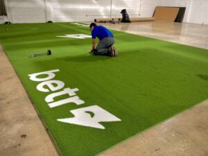 A worker installing artificial turf with a custom logo in a large indoor space by Global Flooring Enterprise in Miami, FL.