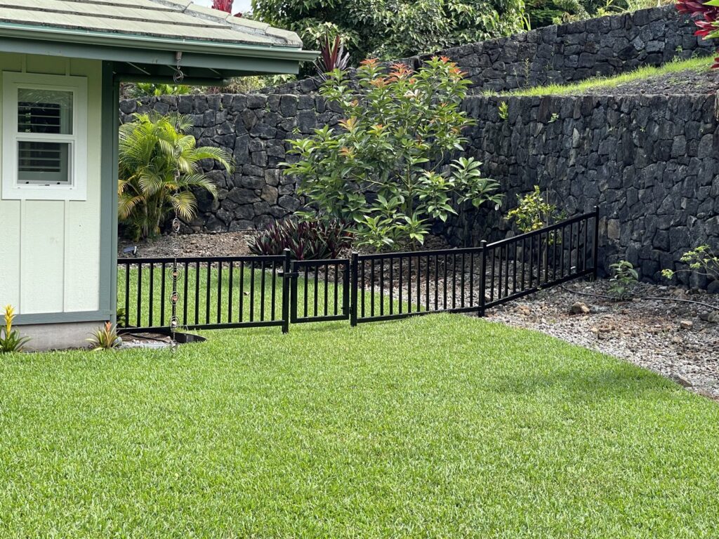 A backyard featuring a new black metal fence with a gate and a rockwall by Fence, Gates and Rockwalls in Kailua-Kona, HI