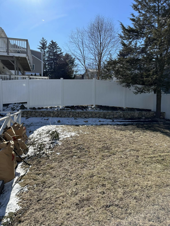 A wide view of a backyard with a newly installed white vinyl fence and stone retaining wall by Hunter & Sons Fence in Burlington, MA.