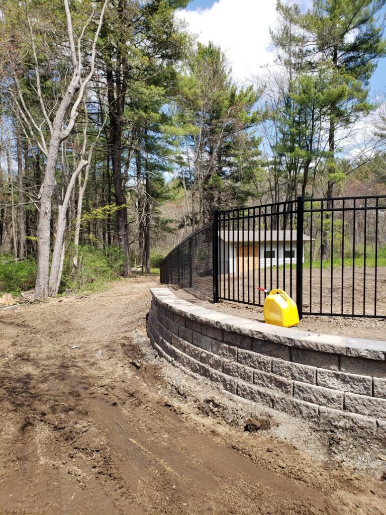 A black ornamental aluminum fence installed on a retaining wall bordering a forest by Sullivan & Sons Fence LLC in Hudson, NH.
