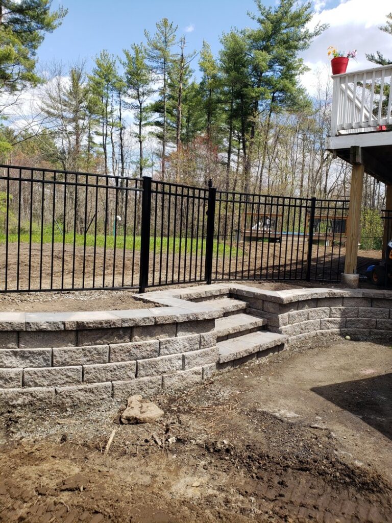 A sleek black ornamental aluminum fence installed atop a stone retaining wall by Sullivan & Sons Fence LLC in Hudson, NH.