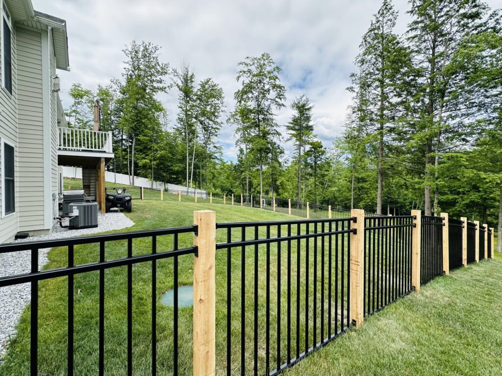 A black aluminum fence with wooden posts installed on a grassy slope by Ultra Fence LLC in Brookline, NH.