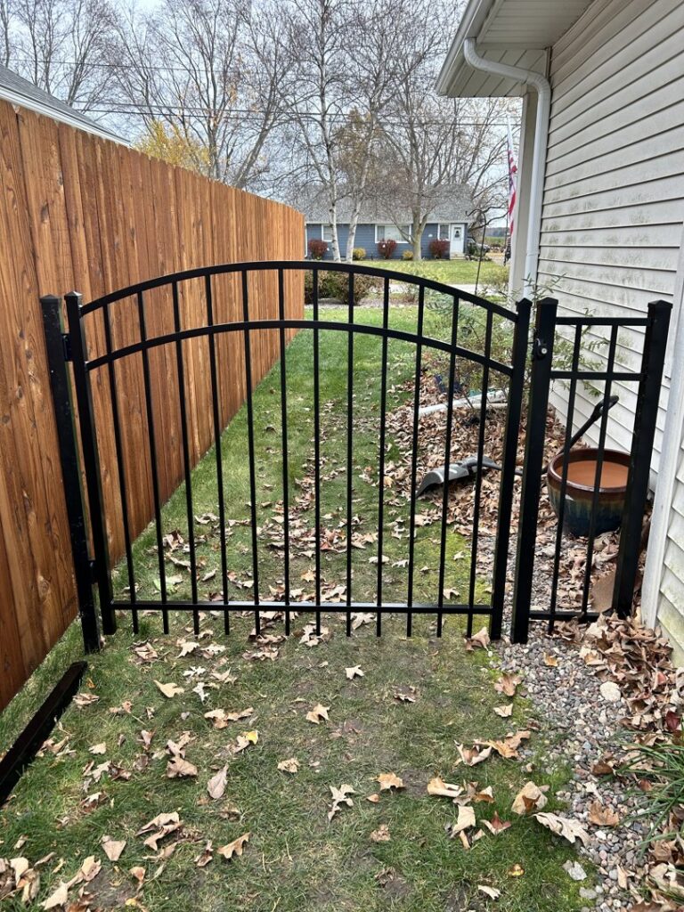 A black ornamental aluminum gate installed next to a wooden privacy fence in a side yard by Safe Yard Fence - Waukesha County in Waukesha, WI.