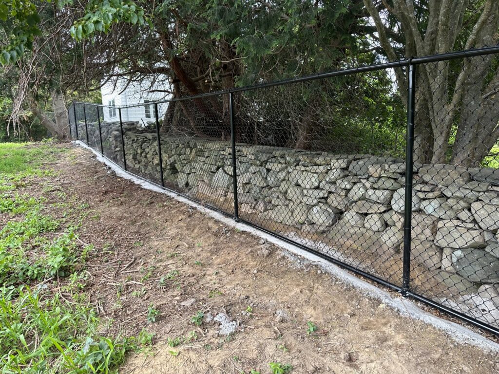 A black chain-link fence installed along a stone wall by Fence Pros Direct in Saunderstown, RI.