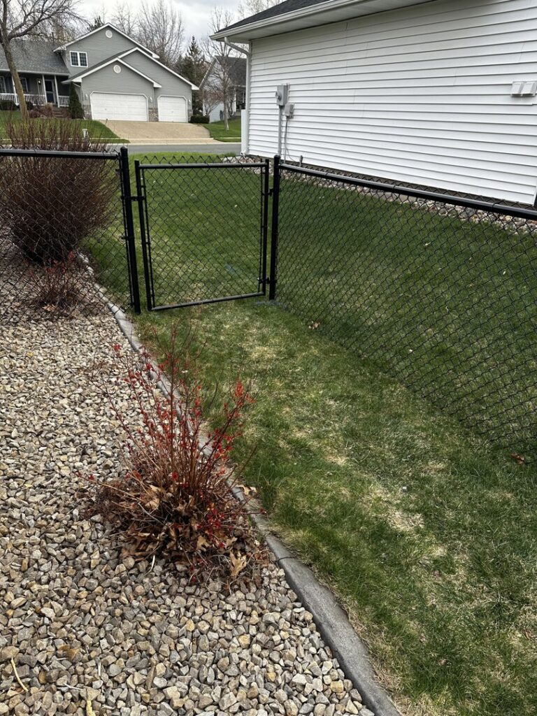 A black chain-link fence with a gate separating a gravel and grassy area, installed by Mauer Company in Little Canada, MN.