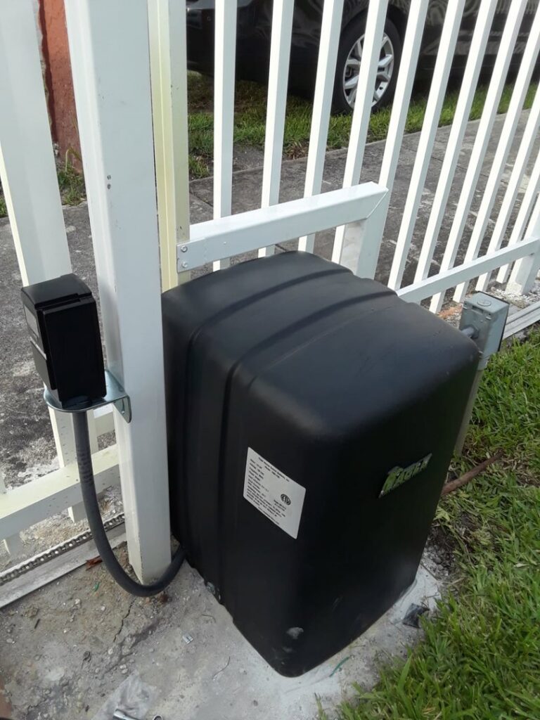 A close-up of a black gate operator motor installed next to a white picket fence by Motor & Fence Solutions in Miami, FL.