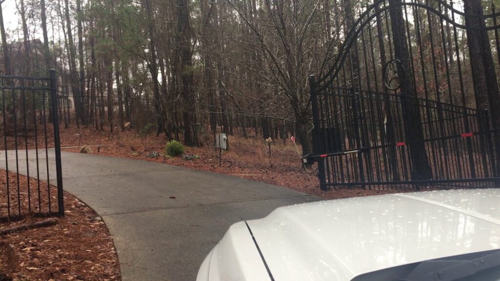 A large black metal driveway gate at the entrance to a wooded property by United Fence Company in Johnston, RI.