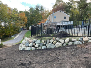 A black metal fence installed atop a decorative rock retaining wall in a landscaped yard by David Platania Fence in Derry, NH.