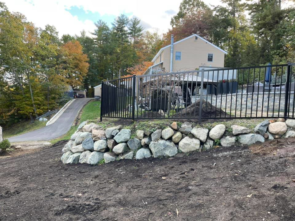 A black metal fence installed atop a decorative rock retaining wall in a landscaped yard by David Platania Fence in Derry, NH.