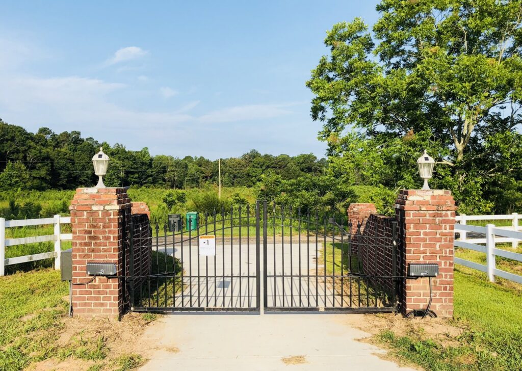 A black metal gate with brick pillars and a white rail fence in the background by Jefcoat Fence Co. Inc. of Hattiesburg, MS
