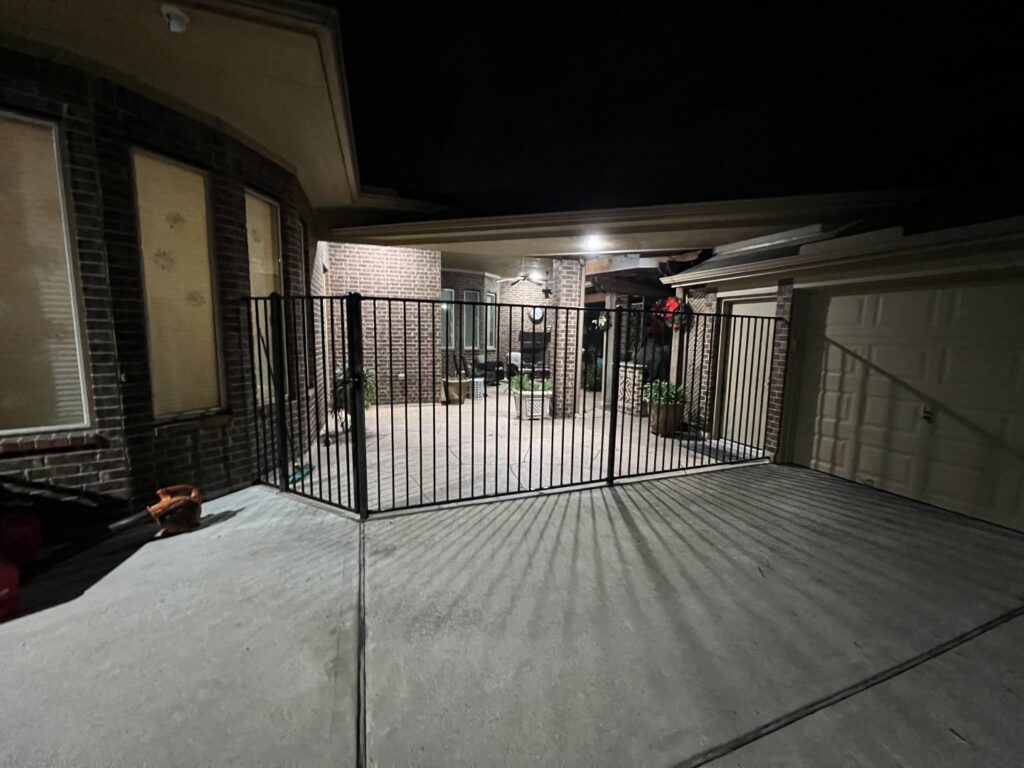 A black metal fence enclosing a large patio and driveway area at night, installed by GOAT Fence Company in Houston, TX.