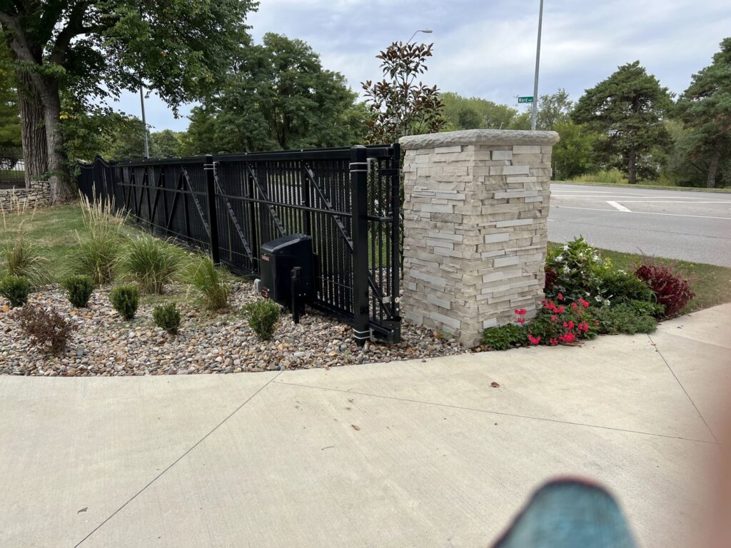 A black metal sliding gate with a decorative stone pillar installed by Brothers Gate Operators and Services LLC in Olathe, KS.