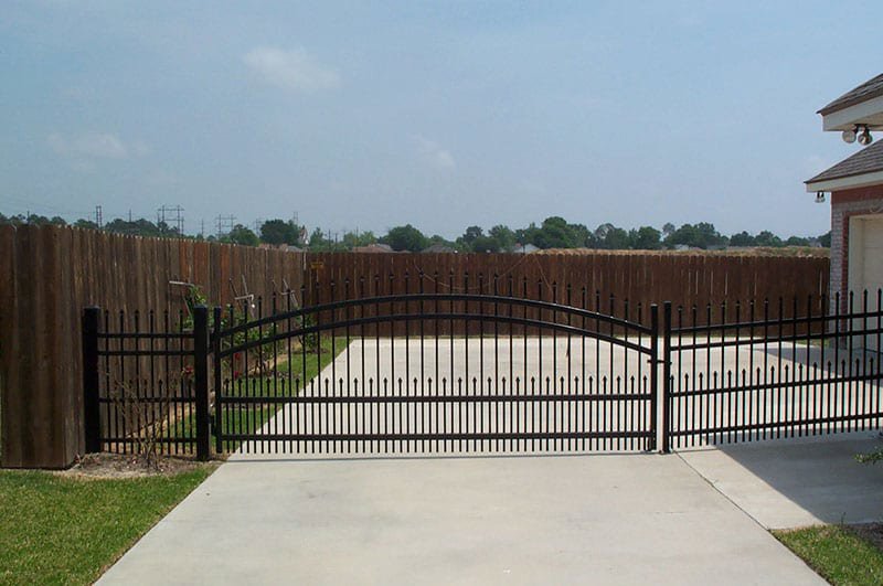 A black ornamental driveway gate with a wooden privacy fence installed by Nash Fence & Supplies Inc. in Lake Charles, LA.