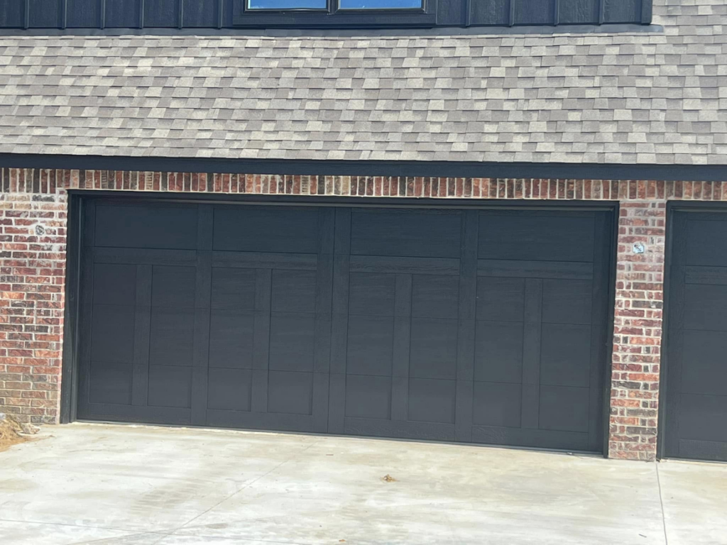 A modern black panel garage door installed on a brick house by L & S Overhead Door in Tulsa, OK.