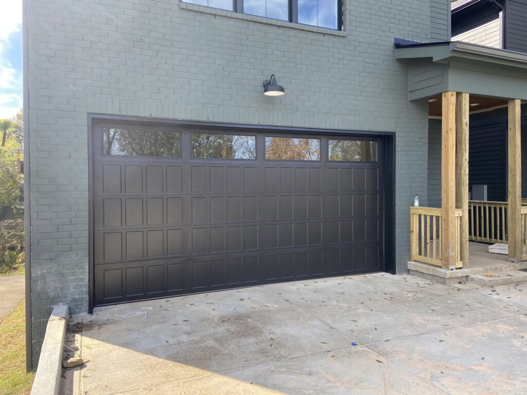 A black residential garage door with top windows installed on a green brick house by Northern Door Company in Murfreesboro, TN