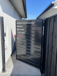 A black slatted gate installed between a house and a dark wooden fence by Bespoke Gate Fence & Deck in Christchurch, NZ.