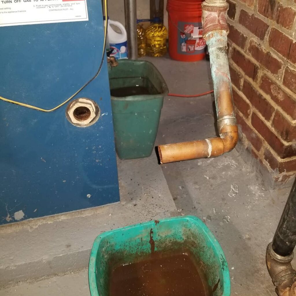 A boiler drain pipe dripping into a bucket, indicating a repair or maintenance job by Batista Heating & Cooling, LLC in Charlotte, NC.