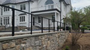A modern black cable railing fence installed along a stone retaining wall next to a large house by H&M Fence, LLC in Norwalk, CT.