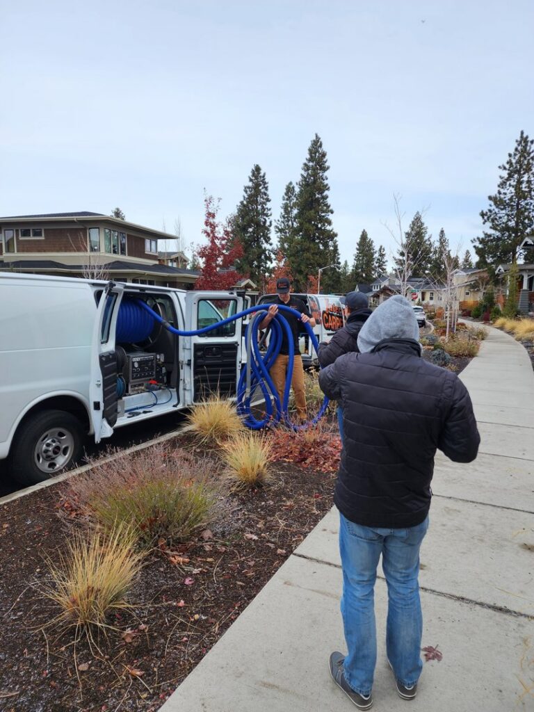 Workers from Interior Care Carpet Cleaning setting up carpet cleaning equipment with blue hoses from a service van in Bend, OR.