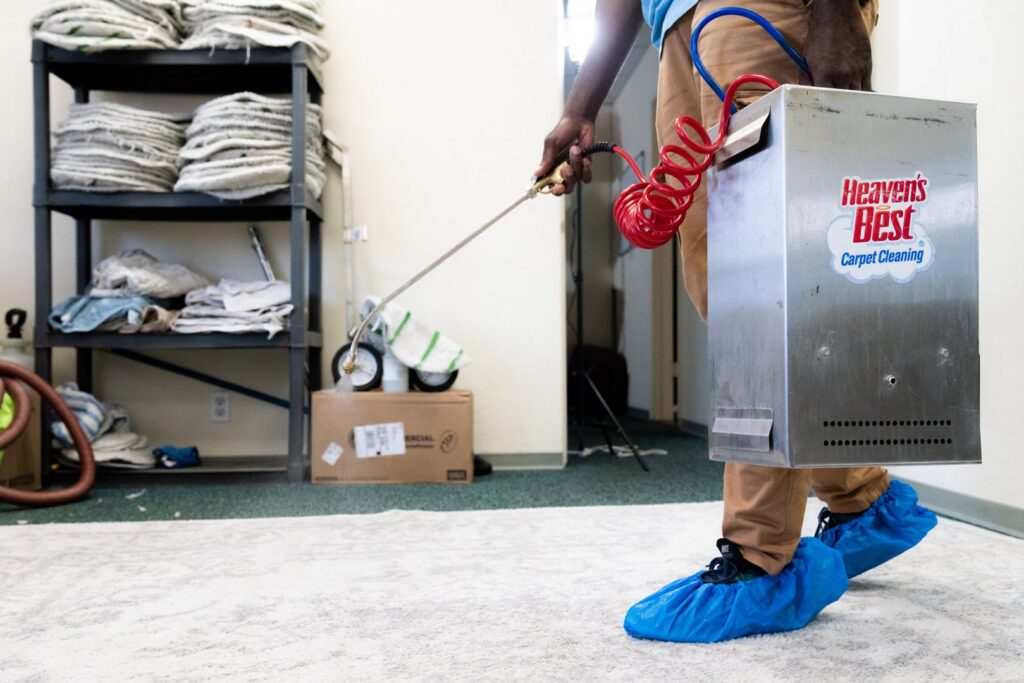 A technician in blue shoe covers using a cleaning wand with a portable unit on a light carpet at Heaven's Best Carpet Cleaning San Diego CA.