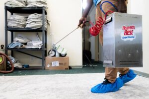 A technician in blue shoe covers using a cleaning wand with a portable unit on a light carpet at Heaven's Best Carpet Cleaning San Diego CA.