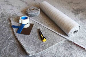 Carpet installation tools including a measuring tape, cutter, and a roll of grey carpet, ready for a job by Gator Carpet and Tile in Jacksonville, FL.