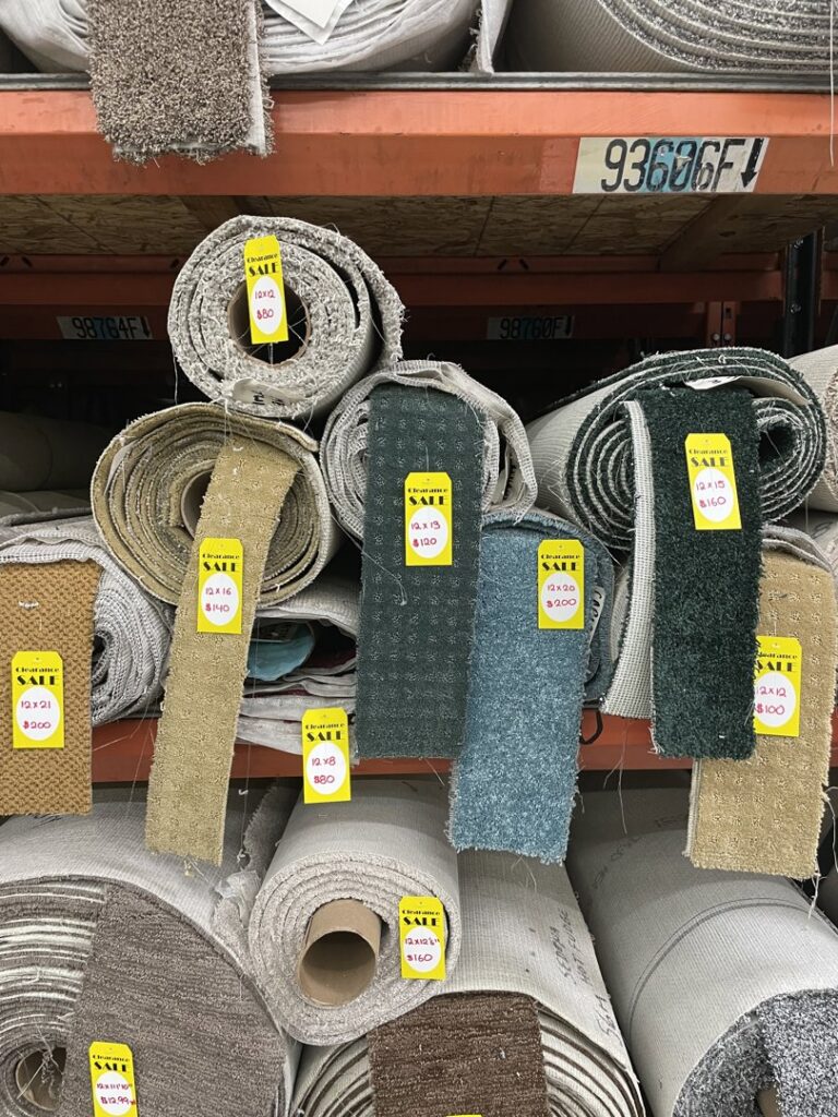 Rolls of various carpets displayed on shelves with 'SALE' tags in the warehouse of Sy's Discount Carpets in Los Angeles, CA
