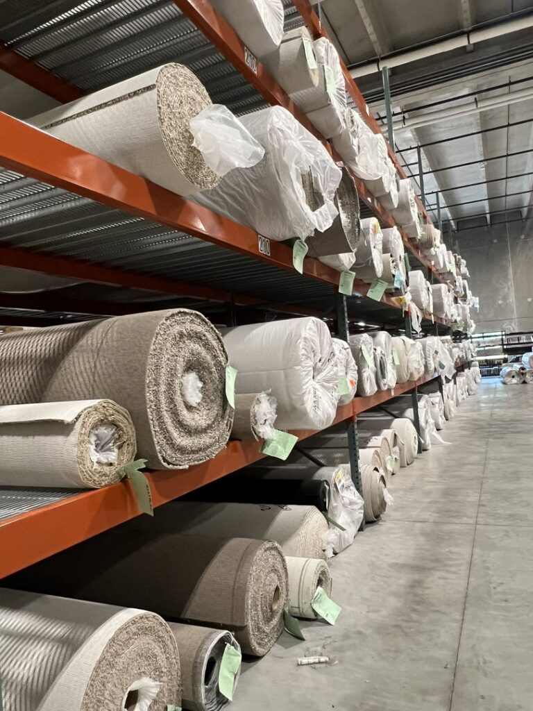 Large rolls of various carpets stored on high shelves in the Eastpointe Interiors warehouse in Caledonia, MI, ready for installation.