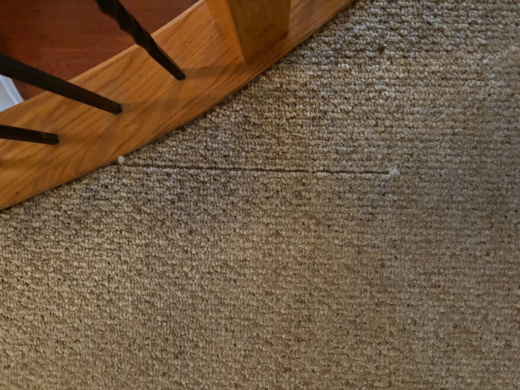 A visible seam or tear in carpet near a wooden staircase, indicating a carpet repair job by Hughes Dry Professional Carpet Care in Marietta, GA.