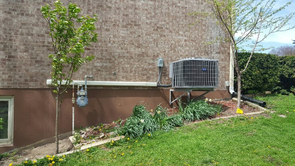 A Carrier air conditioning unit installed on a wall bracket next to a brick house by Custom Climates in Iowa City, IA.