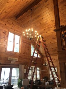 An electrician installing a large chandelier in a high-ceiling room for Bacon Electric, LLC in Concord, NH.