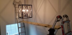 A decorative chandelier being installed in a high ceiling area, with ladders and a plank set up by Maiolo Electric in Pittsburgh, PA.