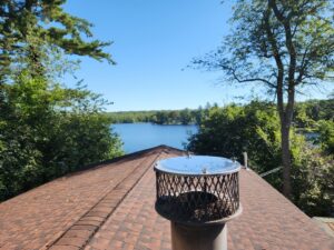 A chimney cap on a residential roof with a scenic lake view, installed by Top Dog Hearth Repair & Maintenance, LLC in Worcester, MA