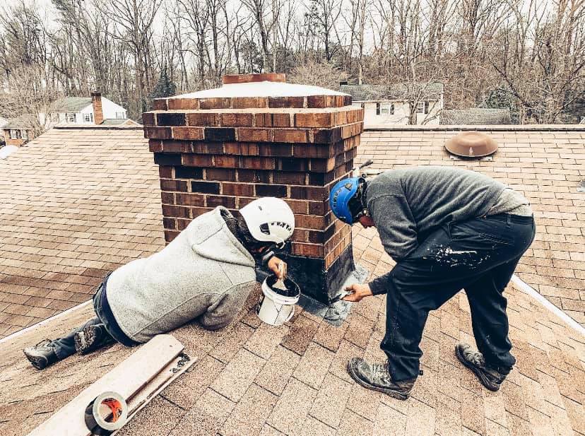 Two chimney sweep technicians repairing and sealing a chimney on a roof for Rooftop Chimney Sweeps in Alexandria, VA.