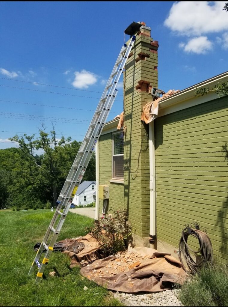 A ladder leaning against a brick chimney undergoing repair, with debris on the ground, by Eagles Nest Chimney Services and more of Cincinnati LLC in Cincinnati, OH.