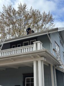 A chimney sweep working on a roof with ladders, performing a chimney service for Chimney Cricket Cleaning Service in Marshfield, MO