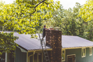 A chimney sweep from Soot Busters on a ladder on a roof, performing work on a brick chimney in Birmingham, AL.