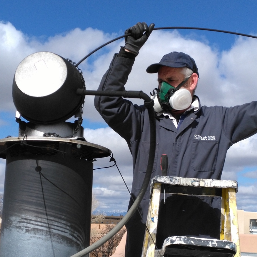 A K-West NM chimney sweep technician on a rooftop, wearing a respirator mask while cleaning a chimney vent in Santa Fe, NM.