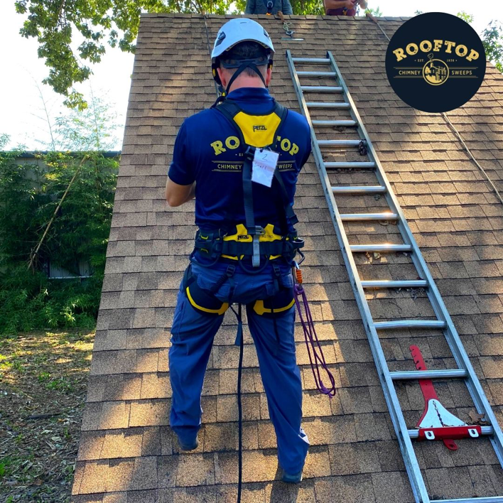 A chimney sweep technician in safety gear on a roof with a ladder for Rooftop Chimney Sweeps in Alexandria, VA.