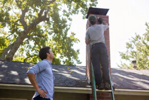 A chimney technician on a ladder working on a brick chimney, observed by a client, for A Sweep Across the Bay in Parrish, FL.