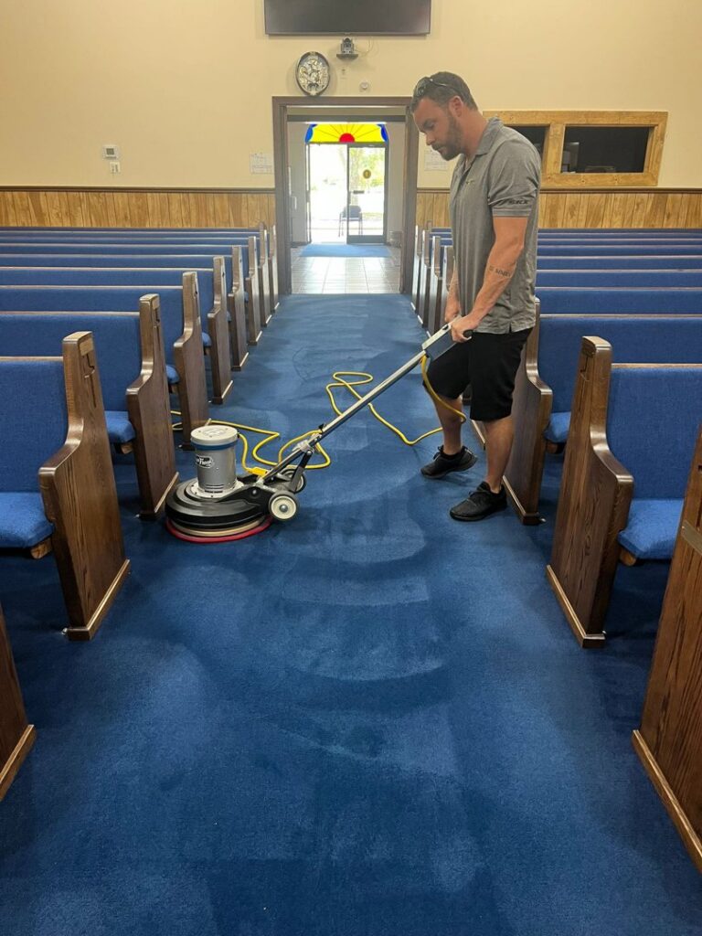 A technician performing professional carpet cleaning in a church aisle with blue carpet by All Ways Organic Citrus Carpet & Upholstery Cleaning in Wilmington, NC.