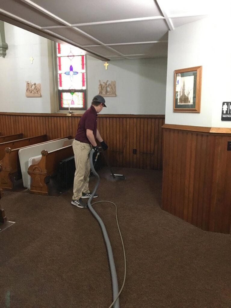 A professional from Yellow Finch Carpet Cleaning LLC cleaning brown carpet in a church, providing commercial services in Dover, NH.