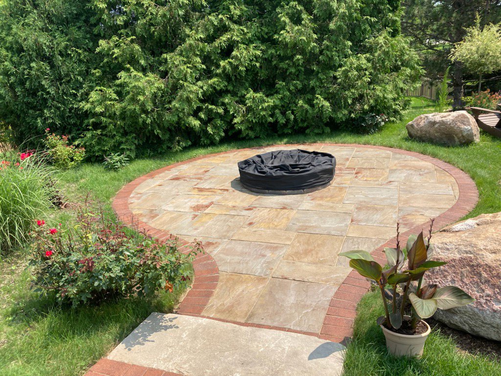 A circular paver patio with a fire pit, surrounded by a lush green lawn and garden beds by Miguel Landscaping LLC in Janesville, WI.
