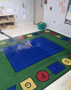 A BeClean technician in Charlotte, NC, steam cleaning a colorful classroom rug, with steam visibly rising.